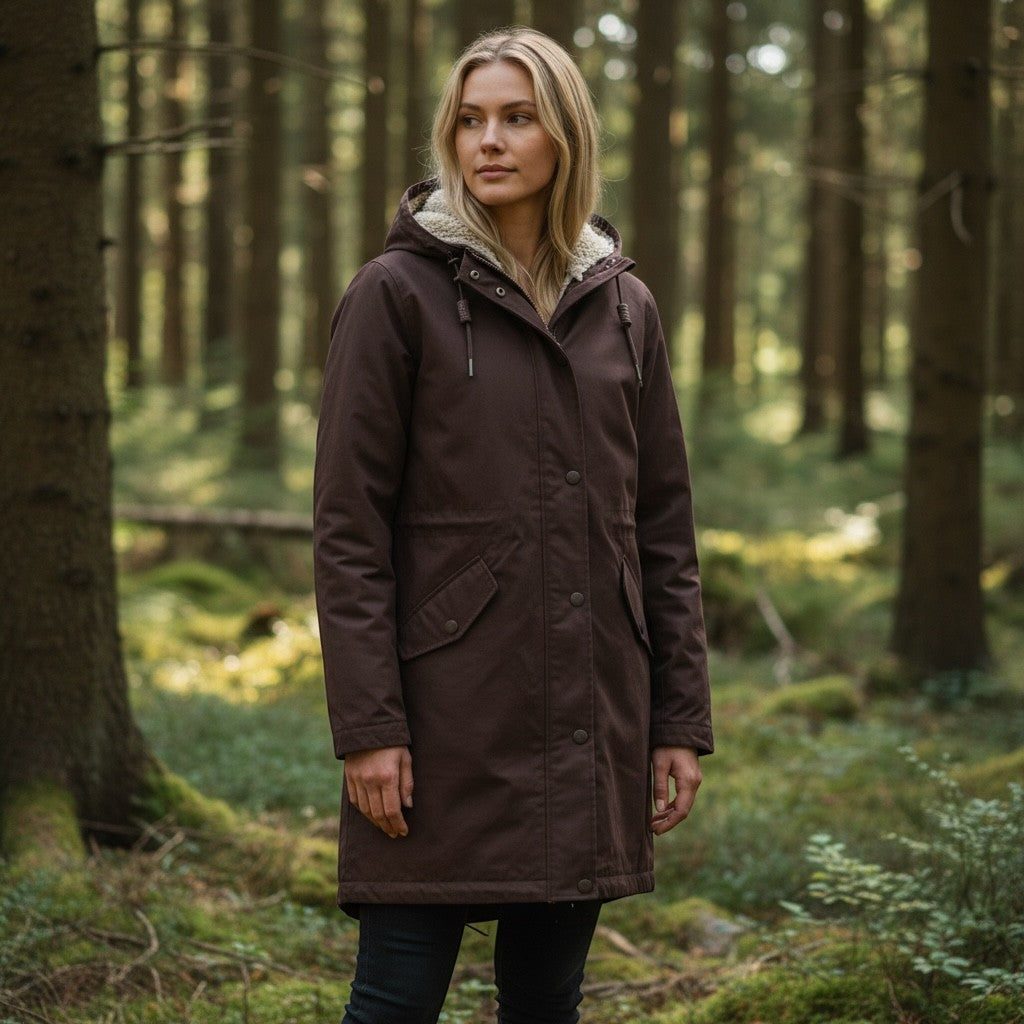Woman wearing a brown Get The High Street Seasalt Plant Hunter waterproof coat with a hood in a forest setting.